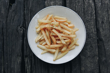 A plate of french fries sits on a wooden table. The fries are seasoned with salt and pepper.
