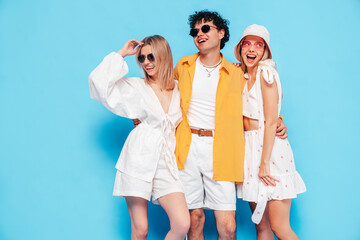 Fototapeta premium Group of young three stylish friends posing in studio. Fashion man and two cute female dressed in casual summer clothes. Smiling models having fun. Cheerful women and guy near blue wall, sunglasses