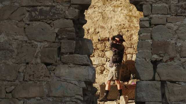 A charismatic gentleman explorer in a hat looks through a lens while standing in an old fortress next to a chest. View through a breach in the ancient fortress wall