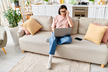 Fototapeta premium A mature woman in cozy homewear sits on a couch, focused on her laptop screen.