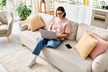 Obraz premium A mature woman in cozy homewear sits on a couch, fully focused on her laptop.