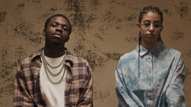 Portrait of stylish African American hip hop couple wearing trendy clothes and accessories sitting down on wooden stools and posing confidently at camera in loft style studio