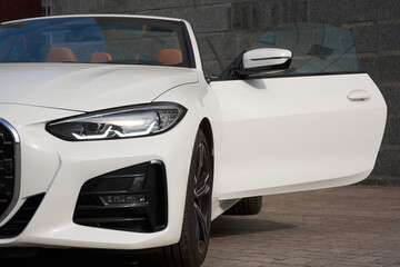 A white convertible car with a brown interior is parked on the street, against a gray wall, with the driver's door open.