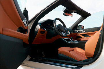 A view of the driver's seat in a white convertible car with a brown interior, as seen through the open driver's door, on the street.