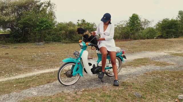 Scenic shot of an Asian woman in a white dress petting a black dog while riding a classic Honda C70 motorcycle. Ideal for travel videos, lifestyle content and dog related content.