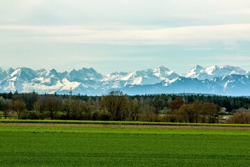 View of the Alpine foothills from the outskirts of Munich. In the foreground are green fields and forests without leaves. Mountains are not in focus