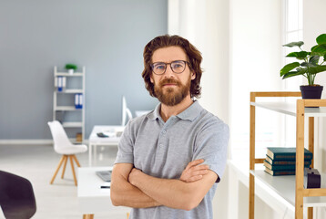 Fototapeta premium Portrait of smart and confident employee who poses against background of modern office. Millennial Caucasian bearded man in casual clothes stands with his arms folded and looks at camera.