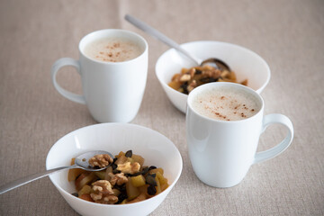 A cozy breakfast setting with two white cups of frothy cappuccino and two bowls of baked apple, nuts and pumpkin seeds on a beige tablecloth, perfect for a relaxed and healthy morning.
