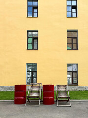 Two chairs and two red barrels are placed against the background of a yellow wall with windows