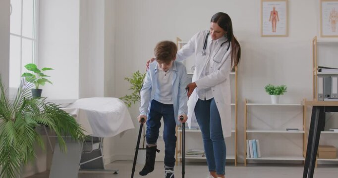 Portrait of a cute child boy with injured bandaged broken leg walking with crutches in medical clinic with a help of young friendly family doctor traumatologist. 4k video. slow motion video.