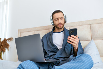 A man enjoys the morning, using a laptop and headphones on a bed.