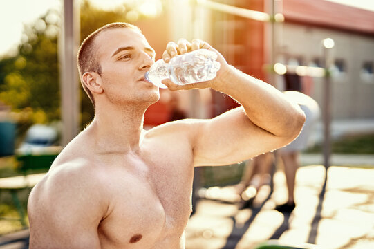 Young muscular athletic sportsman drinking water from a plastic bottle in outdoor fitness park - Hydration and refreshment lifestyle concept with a half-naked millennial bodybuilder