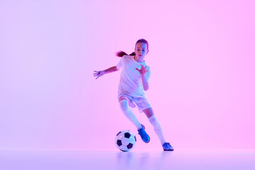 Concentrated teen girl, soccer player training, dribbling ball against pink studio background in...