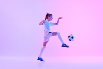 Teen girl in white uniform, soccer plater in motion, practicing, kicking ball against pink studio background in neon light. Concept of sport, active and healthy lifestyle, childhood, school, hobby