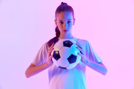 Concentrated football player, teen girl in uniform posing with ball against pink studio background in neon light. Concept of sport, active and healthy lifestyle, childhood, school, hobby