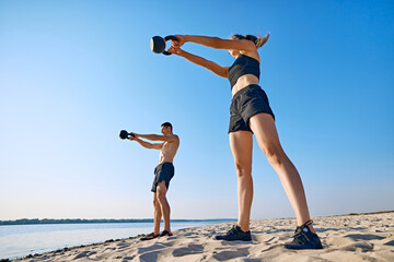 Man and woman with fit and strong bodies performing kettlebell swings on sandy beach, training...