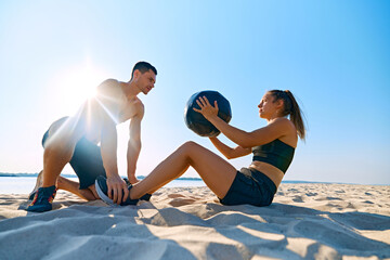 Man helping woman with hard press exercises with medicine ball on sandy beach under clear blue sky. Intensive workout. Concept of sport, active and healthy lifestyle, body care, fitness