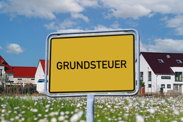 Symbolbild Grundsteuer: Ortsschild mit der Aufschrift GRUNDSTEUER vor einem Neubaugebiet (Composing)