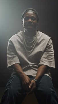 Vertical shot of African American man wearing backwards cap, chain necklace, oversized t-shirt and baggy jeans sitting on chair under projector light in dark room and rapping