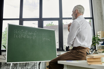 A mature teacher with a beard sits by a whiteboard with math equations, teaching online through a laptop.