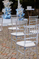 A row of chairs are set up in a courtyard, with a blue and white theme. The chairs are all clear and white, and they are arranged in a neat row. The chairs are empty