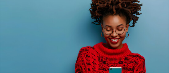 Portrait of smiling african american woman using smartphone, on a blue background, wearing a red sweater and eyeglasses, with space for copy.