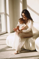 Happy mother in stylish dress sitting on the  sofa and holding her baby daughter in her arms. Mother and daughter in same designed white dresses