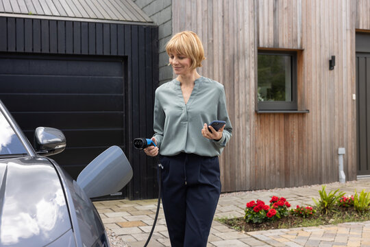 Woman charging electric car outside her home holding her smartphone