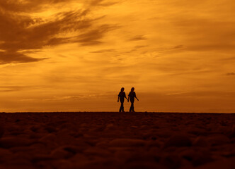 Fototapeta premium female homosexual lesbian Couple walking along coastline during sunset holding hands towards the horizon with cloudy sky and sun as background. couple silhouette walking on beach at sunset in summer