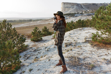 Stylish woman in plaid shirt and hat poses on hill with majestic mountain vista in the distance