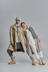 A young, stylish couple stands side by side in trench coats in a studio setting against a grey background.