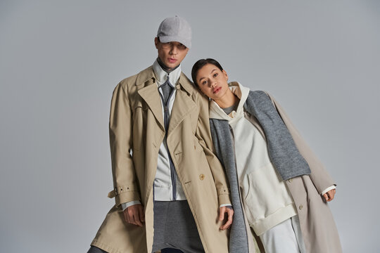 A young, stylish couple stands confidently side by side in trench coats against a grey backdrop.