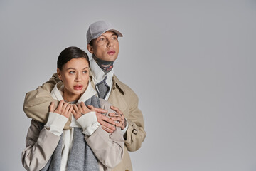 A stylish young man and woman wearing trench coats stand side by side in a studio against a grey background.