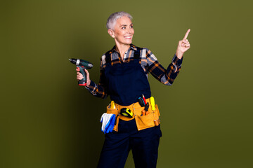 Naklejka premium Photo of mature woman professional mechanic hold drill point look empty space dressed blue workwear isolated on khaki color background