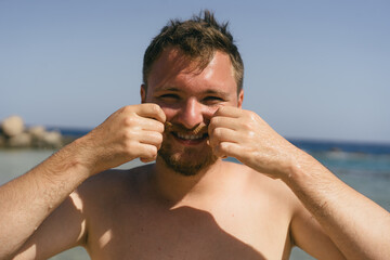 A young man with a beard smiles at the camera while standing in the shallow water on a sunny day at the beach. He is wearing swim trunks and has a relaxed, happy expression.
