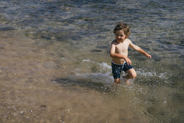 A young boy runs through the shallow water of the ocean on a sunny day. He is wearing blue swim trunks and is splashing water as he runs.