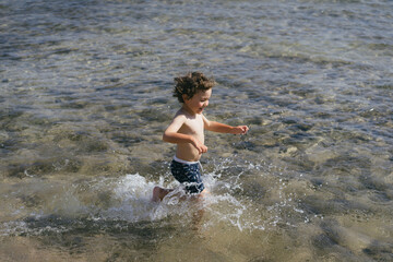 Fototapeta premium A young boy runs through the shallow water of the ocean on a sunny day. He is wearing blue swim trunks and is splashing water as he runs.