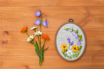 Top view of floral hand embroidery with satin ribbons of summer flowers in hoop. Bouquet of daisies, marigolds and blue bells on wooden background. Crafts and embroidery as hobby. Flat lay, copy space