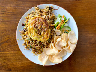 Local Indonesian ketchup fried rice with scrambled egg and prawn crackers, cucumber slices, served on top of white plate, on a wooden table.