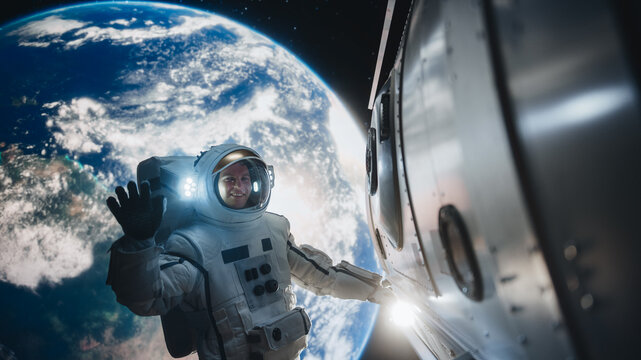 Portrait of a Young Astronaut Posing in a Space Suit During a Spacewalk Outside a Spaceship, Satellite or a Station. Spaceman Looking at Camera, Smiling and Waving Hello Next to an Entrance Hatch
