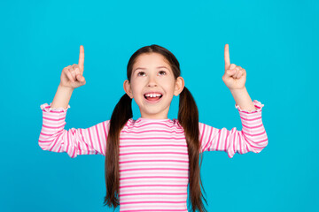 Photo of cheerful small girl with ponytails dressed striped shirt directing look at offer empty space isolated on blue color background