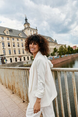 A woman in a white blazer smiles while walking on a bridge in a European city.