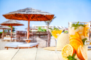 Large pitcher of lemaonade mocktail drink with orange and ice and mint leaves on wooden table with tropical beach background.