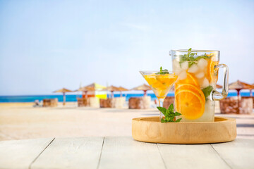 Large pitcher of lemaonade mocktail drink with orange and ice and mint leaves on wooden table with tropical beach background.