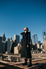A young woman in a black coat dances on a bridge in New York City.