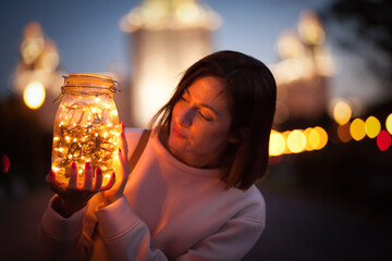 A young beautiful girl holds a jar of lights using it as a lamp in the evening