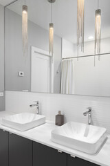 A bathroom detail with pendant lights hanging above vessel sinks and a white marble countertop, a dark grey cabinet, and a view of a tiled shower in the mirror.