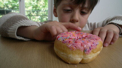 Boy pokes a pink frosted donut with colorful sprinkles on a wooden table, representing childhood desires and indulgence, curiosity and anticipation indoors