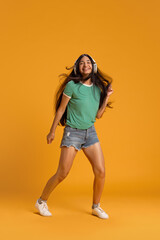 Full-length image of young girl in shorts and t-shirt listening to music in headphones and dancing with joy against yellow studio background. Concept of human emotions, youth, lifestyle