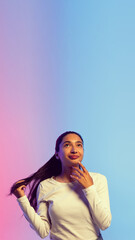 Young woman in white shirt with long black hair, standing with dreamy thoughtful face against gradient pink blue background in neon light. Concept of human emotions, youth, lifestyle